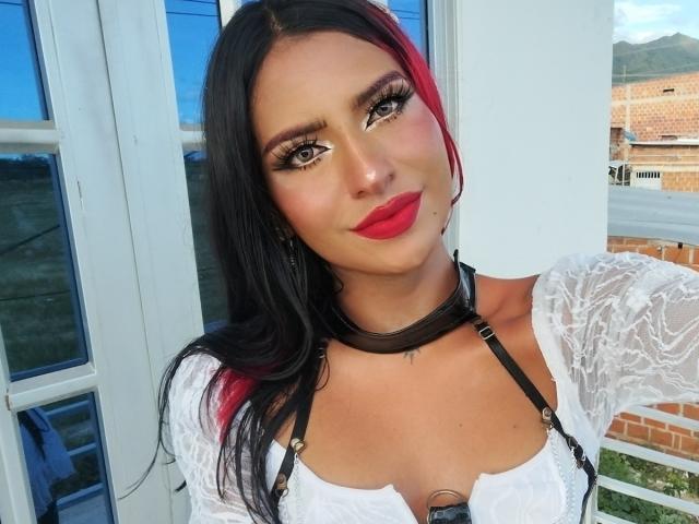 Nefertiti69x ha i capelli neri e rossi, rossetto rosso, e indossa una maglia bianca in pizzo con spalline nere, un choker, e ha un seno abbondante. Sta guardando la fotocamera..