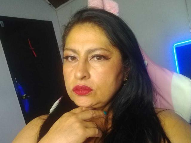 Keyla69, met donker haar, rode lippenstift en dikke eyeliner, die haar lippen opkrult, met haar hand in de buurt van haar kin, een zwarte tanktop, een roze hoofdband en blauwe nagellak, in een kamer..