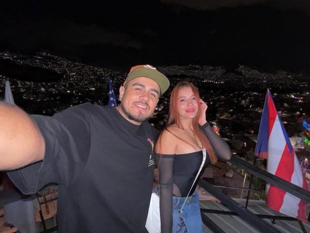 Angeline has tits covered by a black off-shoulder top, standing next to Michael with a chubby ass, wearing a green cap and black shirt, on a rooftop at night.