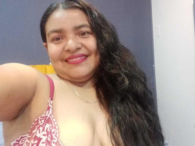 Cataleya Gomez, con lunghi capelli neri e bagnati, sorride, con un reggiseno di pizzo rosa che mette in mostra il suo seno prosperoso, rossetto rosa, collana d'oro, con il braccio sinistro alzato, in un selfie..