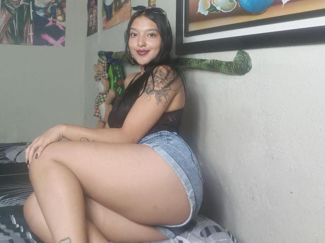 AslyAlexandra, con cabello negro largo, una blusa de encaje marrón, pantalones cortos vaqueros, mostrando su trasero, un tatuaje en el brazo izquierdo, sentada en la cama, sonriendo, una pared con un murciélago verde y obras de arte enmarcadas..