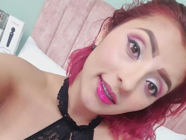 LizBonnet with pink hair, black lace top, pink lipstick, and bold eyeliner, smiling at the camera. Her tits are partially visible, and she's wearing a black choker.