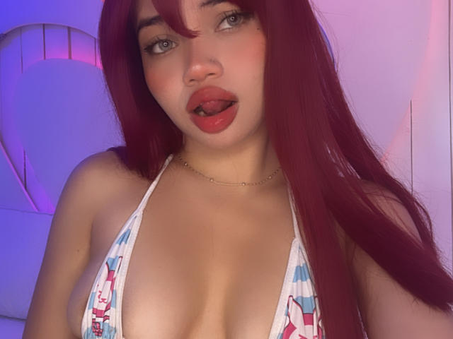 LiliMays with red hair, tongue out, wearing a colorful bikini top, showing her large tits, light skin, and a gold necklace, under purple and blue lighting.