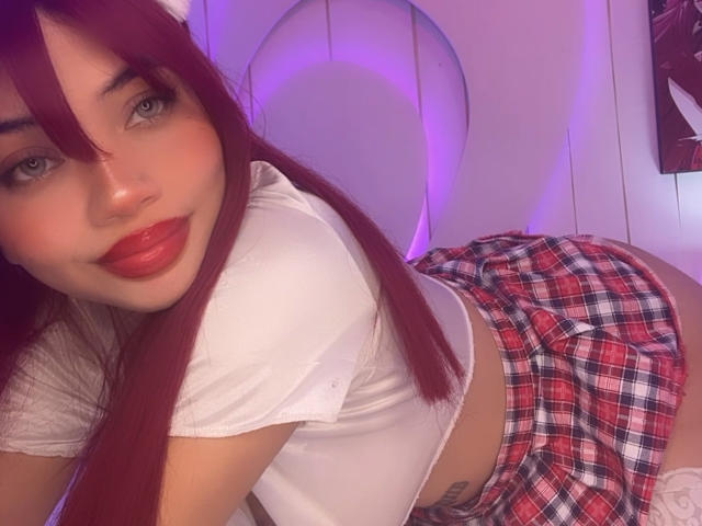 LiliMays with red lipstick, long red hair, white crop top, and red plaid skirt, bent over, showing her ass, in a purple-lit room.