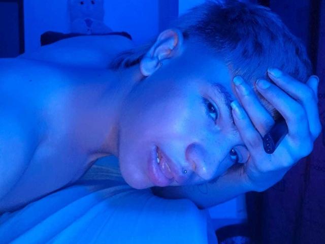 Enrrix met kort blond haar, een huid met blauwige tinten en een lip piercing, ligt plat, met een hand op haar hoofd, kijkt naar de camera, naakt, met een zichtbare tepel in de voorgrond..