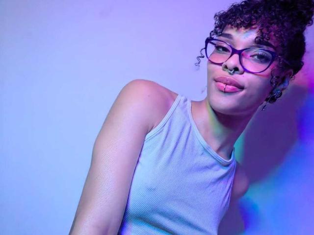 MiaGates, com cabelo castanho encaracolado, óculos, piercing no nariz e piercing no lábio inferior rosa, vestindo uma camiseta cinza, sorrindo levemente. Iluminação azul e roxa..