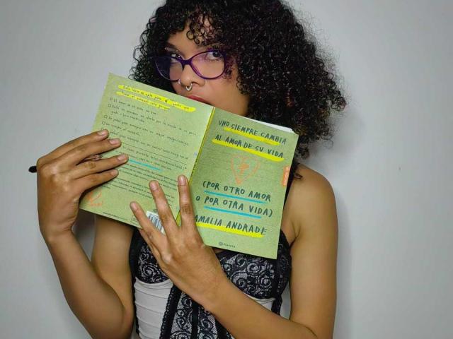 MiaGates, con cabello negro rizado, gafas moradas, piercing en la nariz, sosteniendo un libro verde, vestida con un vestido negro de encaje, mostrando sus senos, mirando el libro, expresión neutra, fondo blanco..