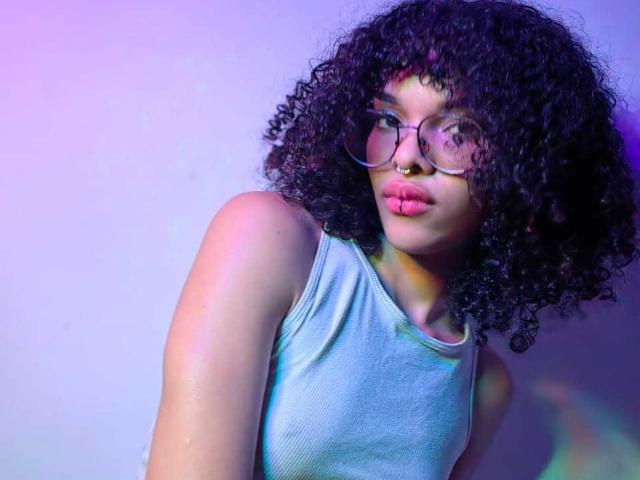 Mia Gates, con cabello negro rizado, usando gafas y un top gris, con labios rosados fruncidos y un piercing en la nariz, en una foto iluminada con luces moradas y rosas..