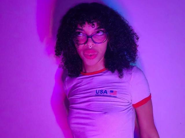 Mia Gates, con cabello negro rizado, gafas, piercing en la nariz y labios rosados, vestida con una camiseta blanca ajustada de Estados Unidos, de pie frente a una iluminación púrpura y azul. Se pueden ver ligeramente sus senos..