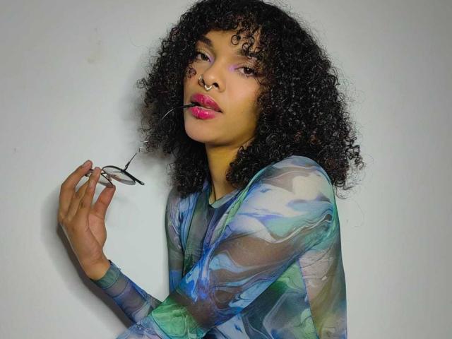 MiaGates, com cabelo preto encaracolado, batom rosa, segurando óculos na mão direita, vestindo uma blusa azul-esverdeada quase transparente, com um piercing no nariz, olhando para a câmera com uma expressão confiante..