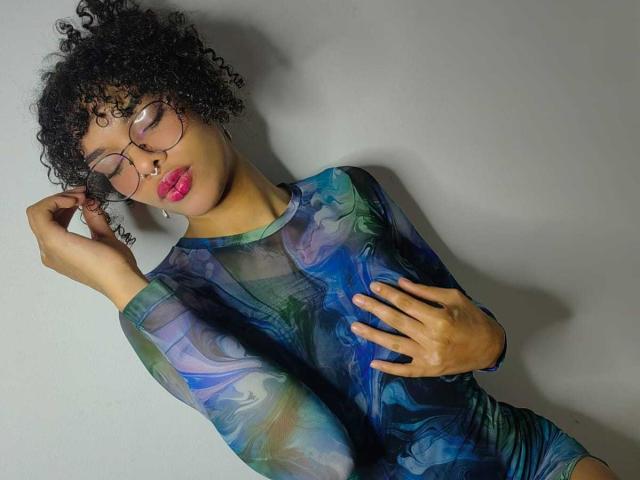 Mia Gates, con cabello negro rizado, gafas y lápiz labial rosa, vestida con una blusa de manga larga de color azul y verde con un diseño marmolado, tocándose el pecho con una mano, de pie frente a una pared gris..
