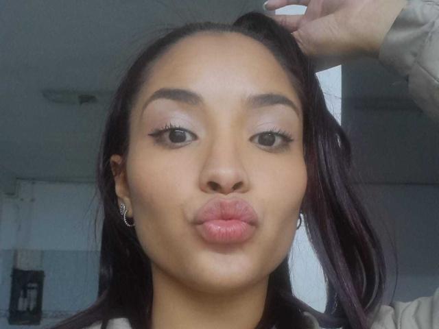 LeaKlarck, avec ses lèvres boudeuses, sa peau brune, ses sourcils foncés et ses longs cils, tenant ses cheveux foncés d'une main, portant de petites boucles d'oreilles, dans une lumière douce..