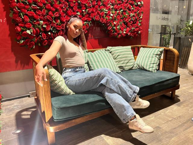 Lea Klarck sitzt auf einem grünen Samtsofa, trägt ein beiges Oberteil, blaue Jeans und weiße Sneakers, und lächelt in die Kamera, während sich im Hintergrund eine Wand mit roten Rosen befindet..