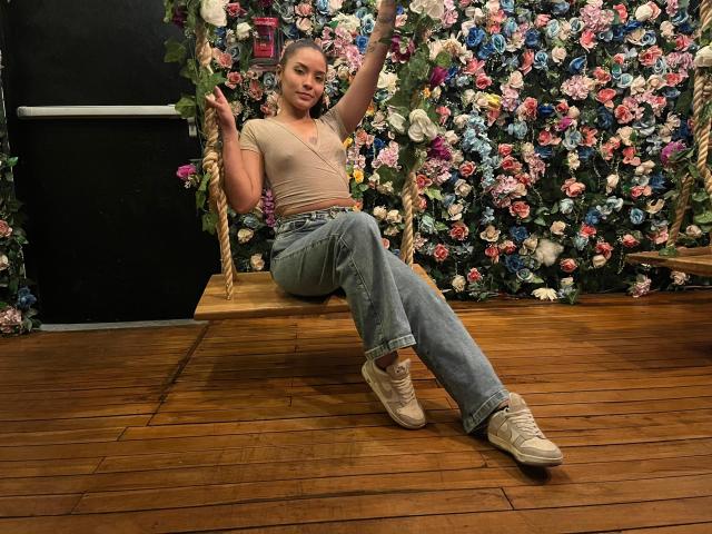 LeaKlarck, con piccoli seni, indossa una maglietta beige e jeans blu, seduta su un'altalena di legno, circondata da rose colorate, con le gambe incrociate e le mani alzate..