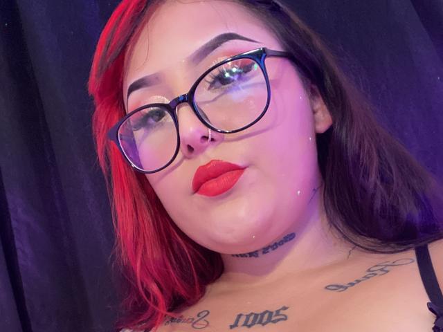 MorganLow mit roten und braunen Haaren, dicken Brillen, rotem Lippenstift, Tattoos auf der Brust und einem schwarzen BH, die in die Kamera lächelt. Strahlende Haut, violettes Licht..