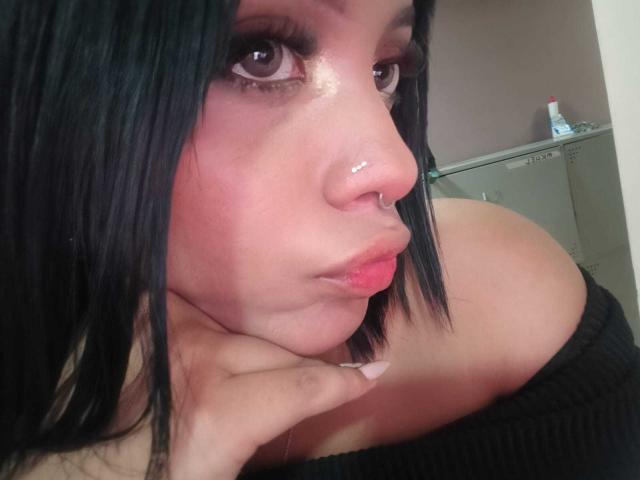 HannaCrow con capelli neri, guance rosate, piercing al naso, labbra rosse, top nero con una spalla scoperta, mano sotto il mento, che guarda verso l'alto. Visibile il seno, illuminazione soffusa..
