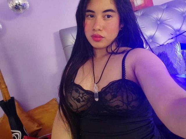 BiancaSolas, con lunghi capelli neri, indossa un reggiseno di pizzo nero, mostrando il seno, e una collana con un ciondolo di cristallo, seduta in una stanza illuminata di viola..