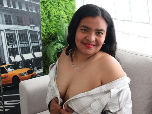 CataleyaGomez with medium brown skin and black hair, smiling, wearing a white plaid shirt pulled down to reveal her large, bare tits. She's sitting on a grey chair.