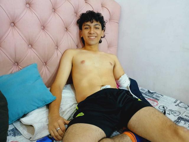 Airlynx, senza maglietta, con capelli neri ricci, sorridente, indossa pantaloncini neri, sdraiata su un letto con una testiera rosa imbottita, un cuscino blu, lenzuola bianche e decorate, e un anello..