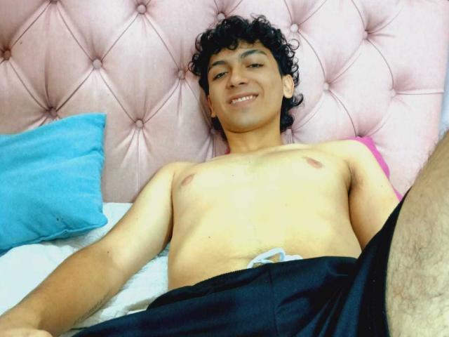 Airlynx est un jeune homme mignon, aux cheveux bouclés, avec un petit pénis, allongé sur un lit, souriant, torse nu, avec un pantalon noir et un oreiller rose derrière lui..