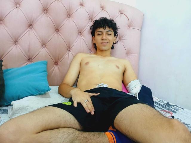 Shirtless Airlynx with curly black hair and light skin, lying on a bed with a pink tufted headboard, wearing black shorts, right hand on crotch, smiling.