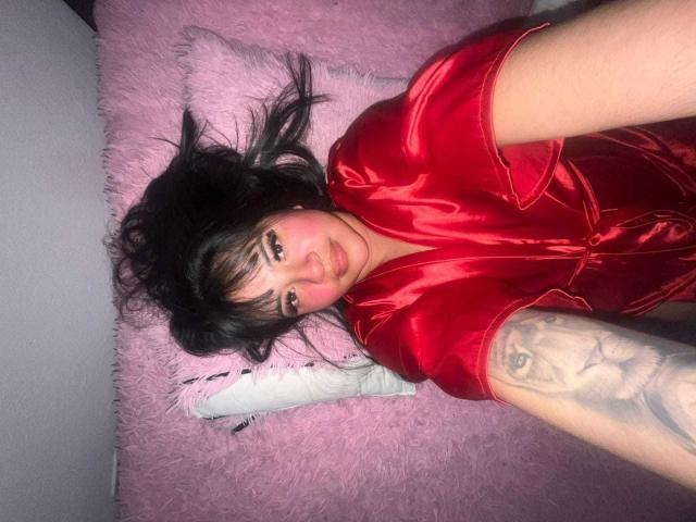 Sofia Rutso liegt auf einem rosafarbenen, flauschigen Kissen, trägt ein glänzendes, rotes Seidenkleid, hat schwarzes, welliges Haar, blaue Augen, ein Tattoo am rechten Arm und lächelt in die Kamera. Halbkörperaufnahme..
