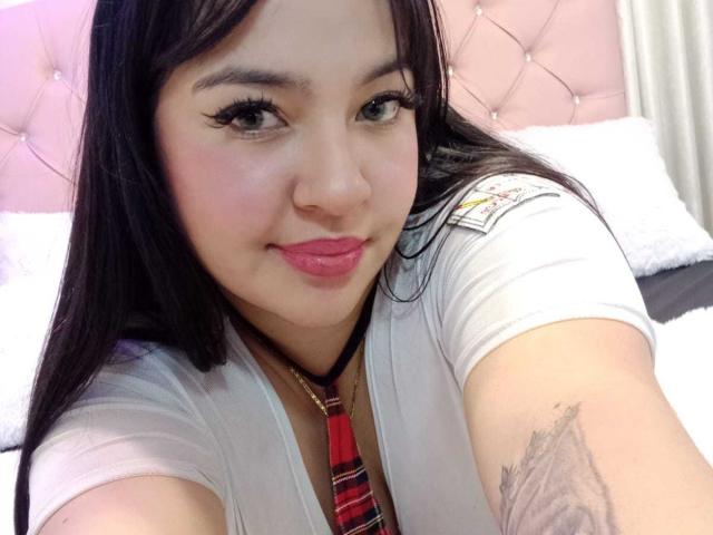 Sofia Rutso mit langem, schwarzem Haar, rosafarbenen Lippen und Eyeliner, trägt eine weiße Bluse mit einem roten Karokrawattenhemd. Auf ihrem rechten Arm ist ein Tattoo sichtbar. Sie lächelt in die Kamera..