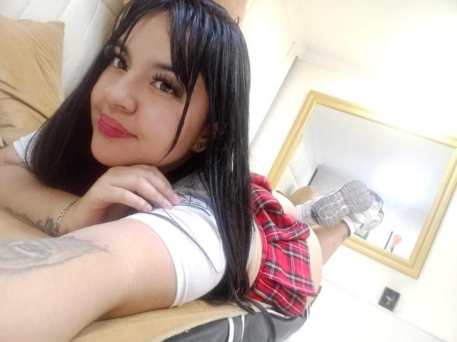 Sofia Rutso, con cabello negro, falda escocesa roja, blusa blanca, acostada en la cama, con las piernas levantadas, mostrando su trasero, lápiz labial rosa, tatuaje en el brazo, en un selfie en el espejo..