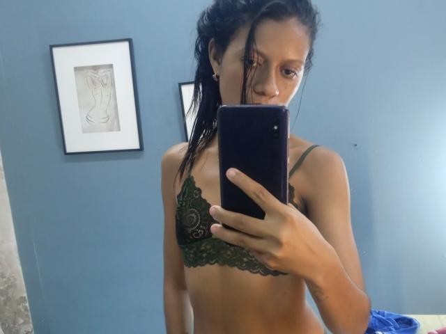MonnicaBella, avec ses cheveux noirs mouillés, porte un soutien-gorge en dentelle vert et prend un selfie devant un miroir, montrant ses petites seins et son corps mince et tonique. Son téléphone est dans sa main droite..