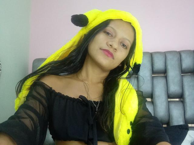 MonnicaBella, avec de longs cheveux noirs, portant un chapeau en forme d'animal jaune et duveteux et un haut noir transparent à épaules dénudées, sourit légèrement. Elle a du rouge à lèvres rose et un collier en argent..