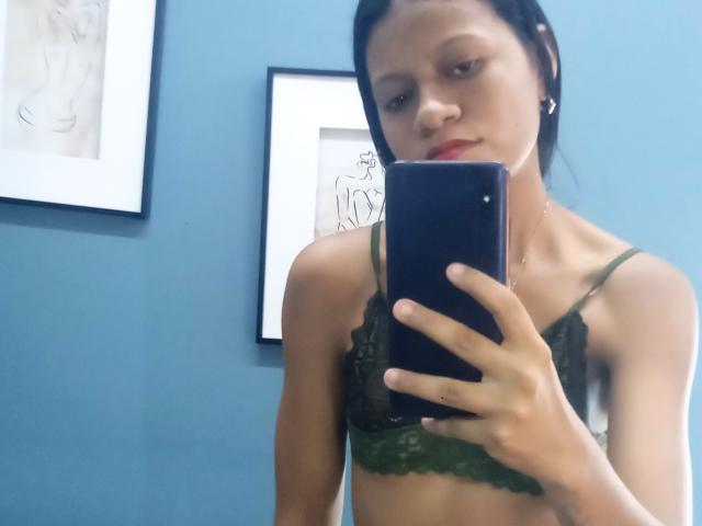 MonnicaBella se toma un selfie frente al espejo con su sujetador de encaje verde puesto, sosteniendo un teléfono azul, mostrando sus pechos, y tiene una sonrisa sutil. Tiene la piel clara y el cabello oscuro..