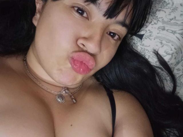 Sofia Rutso, con labbra rosa carnose, capelli neri e occhi marroni, indossa una collana nera a doppia catena con un laccetto e mostra il suo seno prosperoso. È sdraiata e ha un'espressione leggermente accigliata..