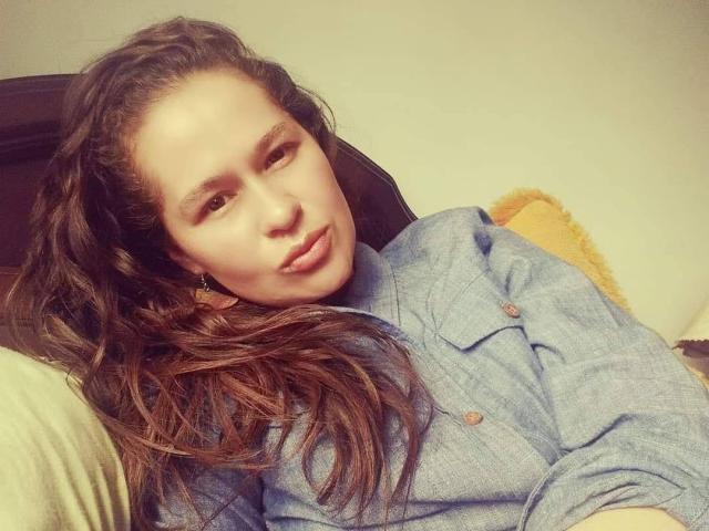 Samantha69, mit welligem, braunem Haar, trägt ein blaues Hemd mit Knopfleiste und hat einen entspannten, leicht schmollenden Gesichtsausdruck. Sie sitzt mit verschränkten Armen in einer gemütlichen Innenraumumgebung..