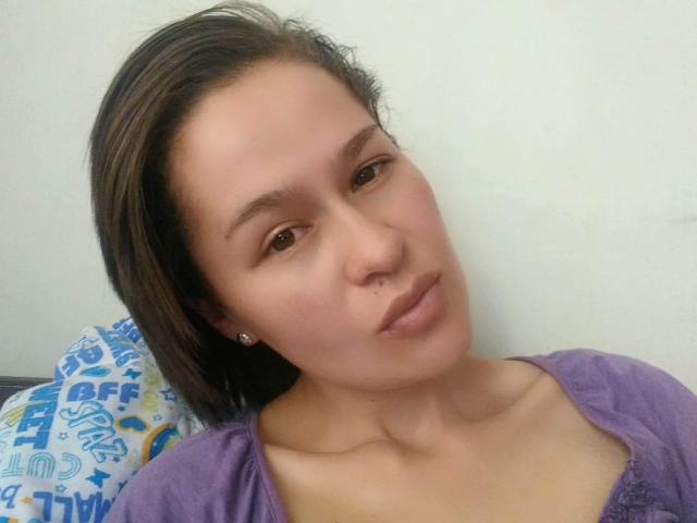 Samantha69Love ha la pelle marrone chiaro, i capelli castani lisci e indossa una maglietta viola. Guarda la fotocamera con un'espressione neutra, e si vedono dei piccoli orecchini..