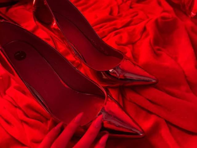 Saltos altos vermelhos com bicos pontudos e brilhantes e solas vermelhas estão colocados sobre um tecido de cetim vermelho. As solas têm um logotipo circular preto. Sapatos sensuais da MiraGi..