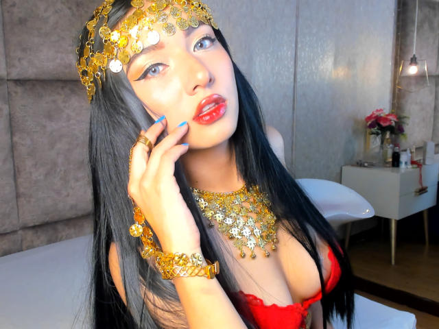 LillyTaylor with long black hair, red lipstick, gold headpiece, and necklace, touching her face, wearing a red dress, showing her boobs, in a bright room.