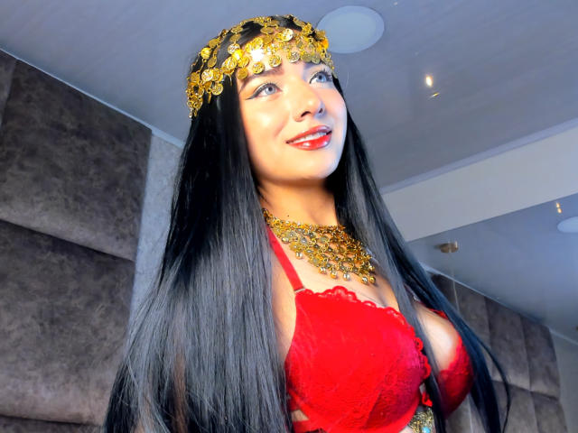 LillyTaylor with long black hair, wearing a gold headpiece, red lace bra, and gold necklace, smiling with red lipstick, showing her big boobs, in a well-lit room.