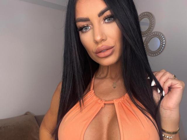 Laylak ha lunghi capelli neri, occhi azzurri e labbra piene, e indossa un top arancione che mette in mostra il suo seno grande, lucido e prosperoso. Sta toccando i capelli con le unghie curate..