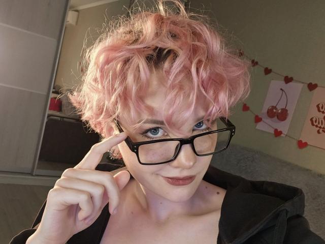 ShellyLove69, com cabelo cacheado rosa, óculos pretos e um leve sorriso, apontando para os óculos com o dedo. Ela está vestindo um moletom preto, mostrando seus ombros nus..