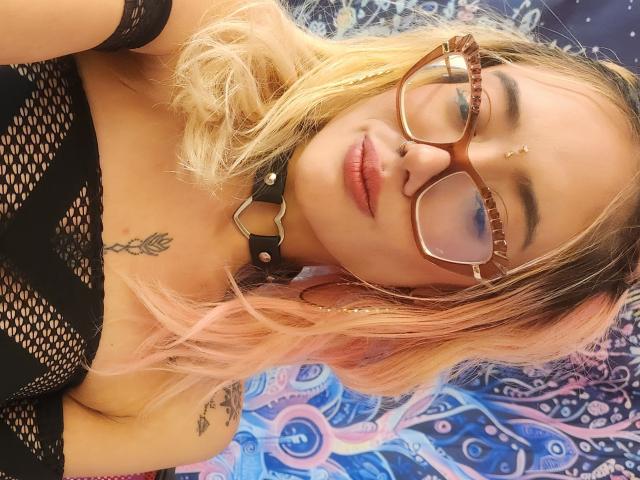 FreankyQueen with blonde and pink ombre hair, glasses, nose ring, and black mesh top, showing tits, wearing a black collar, tattoo on chest, and eyeliner.