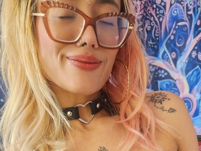 FreankyQueen, avec des cheveux roses et blonds, des lunettes marron, un anneau au nez et des boucles d'oreilles créoles, portant un collier noir avec un pendentif en métal en forme de cœur, souriant avec des lèvres roses..