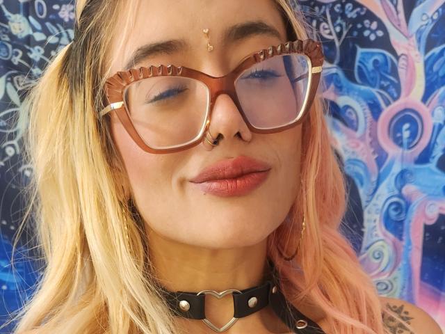 FreankyQueen with pink and blonde hair, brown glasses, nose ring, and heart choker, has a small gold dot on her forehead and pink lips, looking slightly smug.