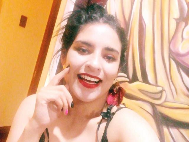 BadAngel con cabello castaño largo, gafas y un traje de baño blanco ajustado, sentada en una cama rosa, con las manos sobre los muslos, mostrando senos prominentes y un ligero vientre, con un fondo de cortina verde..