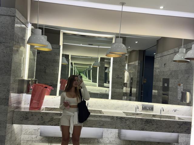 NatSorian, avec de longs cheveux bruns, un débardeur blanc et un short blanc, tenant un téléphone, un sac rose et un sac noir, se tient devant un miroir de salle de bain en marbre, montrant ses fesses..