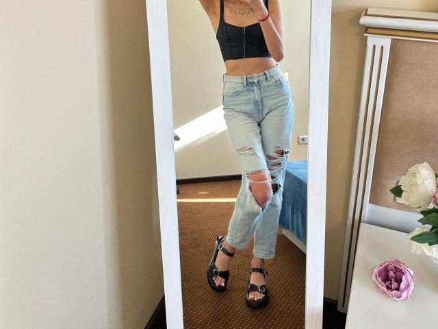 Karish indossa una canotta nera, jeans azzurri strappati, sandali neri con cinturini e ha un tatuaggio sul petto. Ha i glutei piccoli ed è in piedi davanti a uno specchio..