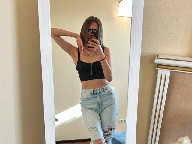 Karish with small tits, black strapless top, light blue ripped jeans, holding phone covering face, mirror reflection, arm raised, tattoo on chest, red bracelet, beige wall.