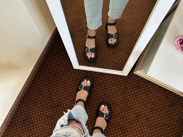 Karish's feet in black sandals with buckles, reflecting in a mirror. Karish wears ripped blue jeans, standing on brown carpet with white trim, pink hair tie visible.