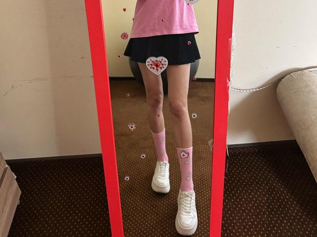 Karish has a pink shirt, black skirt, pink socks with hearts, and white sneakers, reflected in a red-framed mirror. Her ass is covered by a heart sticker.