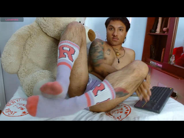 BillyReynols, ohne Hemd, mit einem tätowierten Arm, trägt weiße Socken mit einem rosa "R" und rosa Sohlen, sitzt auf einem Bett, die Beine hoch, mit einem Laptop in der Hand, neben einem riesigen Teddybären..