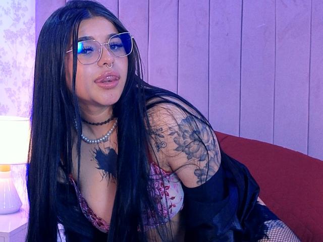 Adhara Evans mit langen, schwarzen Haaren, Brille, Nasenring und Tattoos, trägt eine schwarze Jacke, eine Perlenkette und einen BH mit Blumenmuster, lehnt sich an einen roten Stuhl und hat leicht geschürzte Lippen..